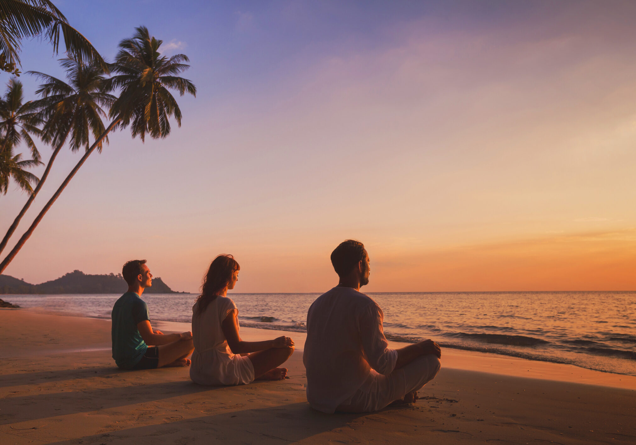 Three people meditating on beach at sunset